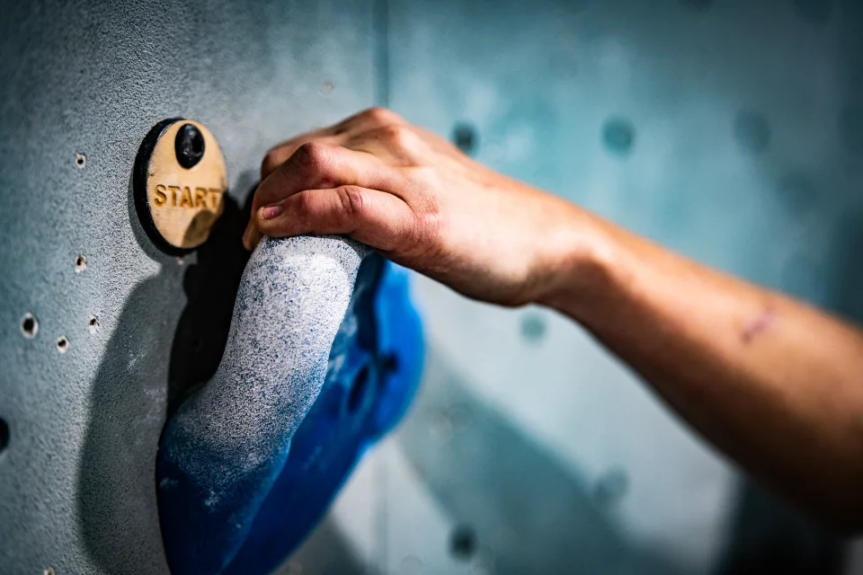 A hand gripping a blue indoor climbing hold beneath a Start marker