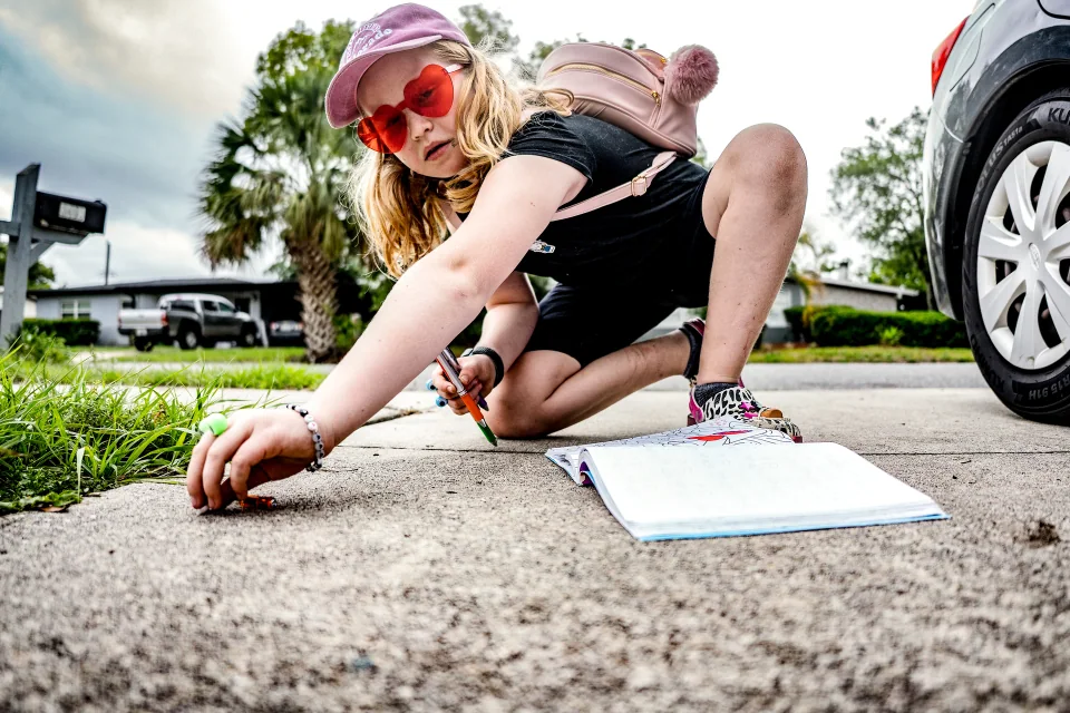 A child crouching on a driveway to draw with chalk beside a parked car