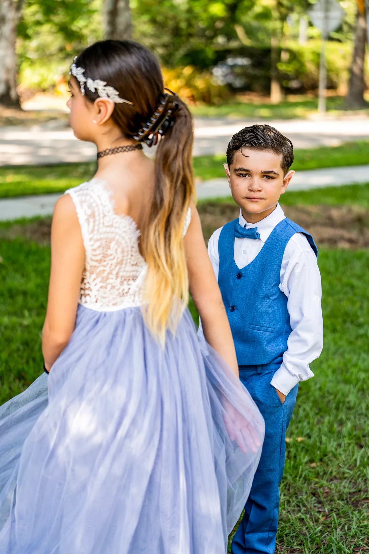 A wedding attendant in a pale dress standing with a child in a blue vest on a garden path