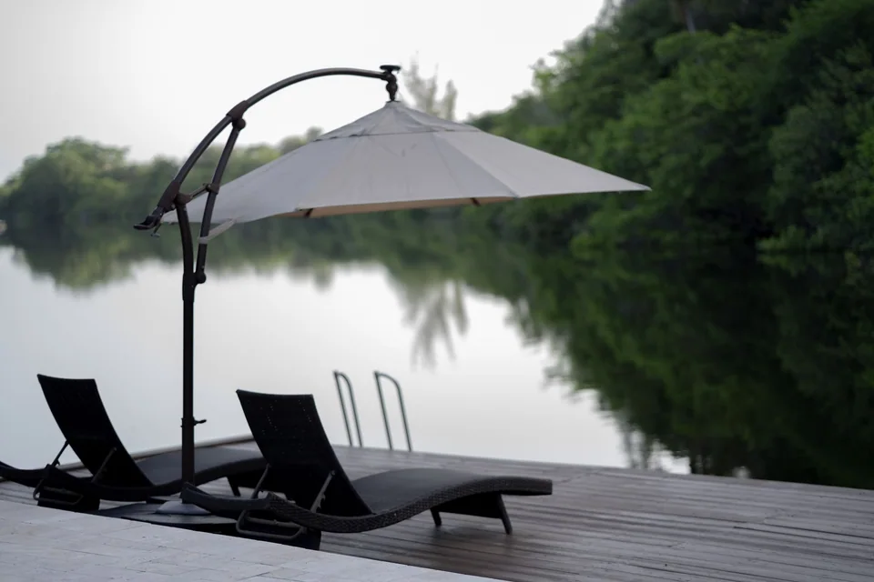 Lounge chairs and an umbrella beside a still lake with trees reflected in the water